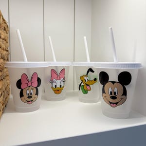 Puede incluir: Cuatro vasos de pl&aacute;stico transparente con tapas y pajitas blancas, cada uno con un personaje diferente de Disney: Minnie Mouse, Daisy Duck, Pluto y Mickey Mouse. Los vasos est&aacute;n colocados en un estante blanco.