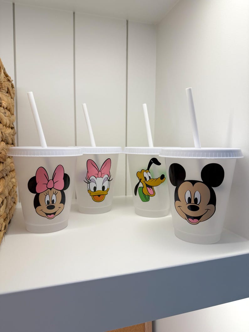 Puede incluir: Cuatro vasos de pl&aacute;stico transparente con tapas y pajitas blancas, cada uno con un personaje diferente de Disney: Minnie Mouse, Daisy Duck, Pluto y Mickey Mouse. Los vasos est&aacute;n colocados en un estante blanco.
