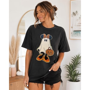 Puede incluir: Una camiseta gris carbón de manga corta con el gráfico de un personaje fantasma que lleva un lazo con estampado de leopardo y zapatos naranjas, sosteniendo una calabaza. El fantasma tiene orejas negras de ratón y un cuerpo blanco.
