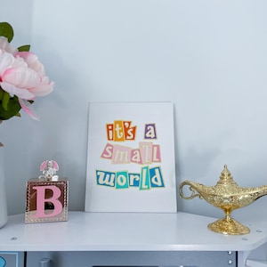 May include: A white framed print with the text "it's a small world" in colorful letters. The letters are cut out and layered on top of each other. The print is sitting on a white shelf with a pink block letter B and a gold genie lamp.