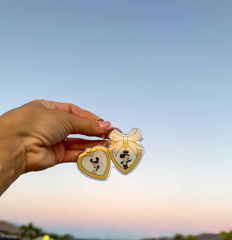 May include: A gold-colored heart-shaped keychain featuring Mickey and Minnie Mouse in wedding attire. The keychain has a bow design and a gold border. The background is a clear blue sky.