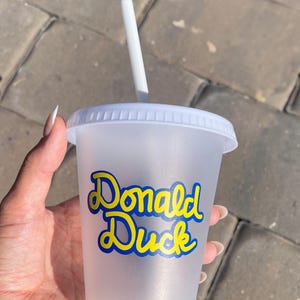 Puede incluir: Un vaso de pl&aacute;stico transparente con tapa y pajita blancas. El vaso presenta las palabras "Donald Duck" en escritura azul y amarilla. El vaso se sostiene en una mano sobre un fondo de ladrillos grises.