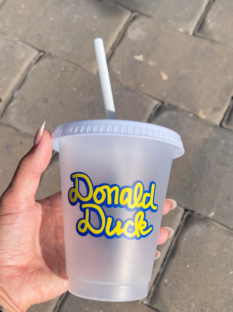 Puede incluir: Un vaso de pl&aacute;stico transparente con tapa y pajita blancas. El vaso presenta las palabras "Donald Duck" en escritura azul y amarilla. El vaso se sostiene en una mano sobre un fondo de ladrillos grises.