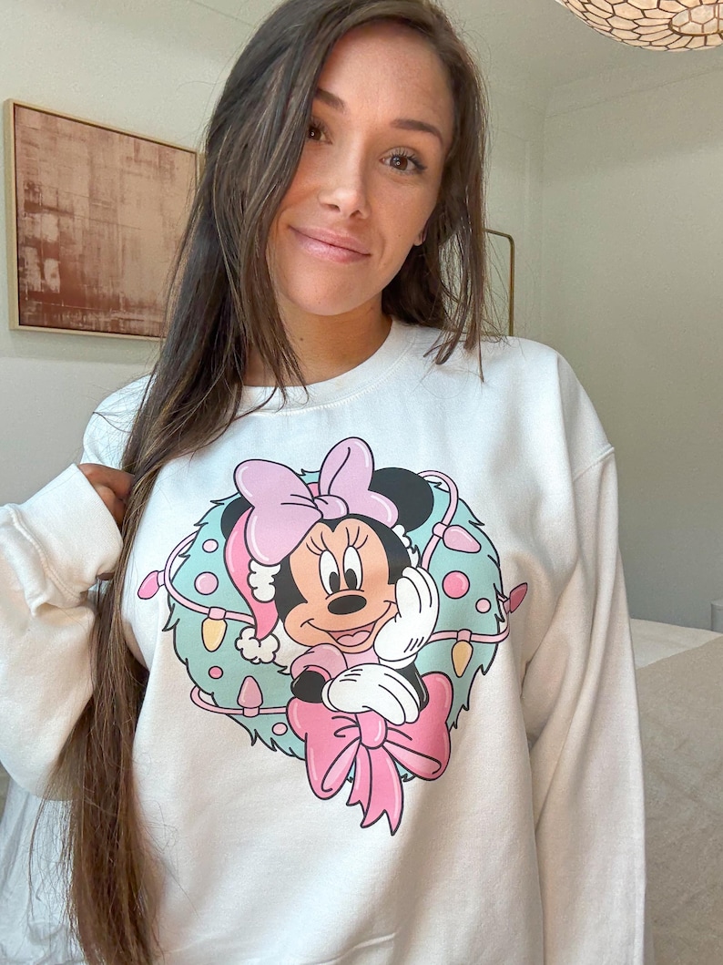 Peut inclure: Sweat-shirt blanc avec un motif de Minnie Mouse. Le motif repr&eacute;sente Minnie Mouse avec un n&oelig;ud rose et un bonnet de P&egrave;re No&euml;l, entour&eacute;e d'une couronne avec des guirlandes lumineuses. Le sweat-shirt a des manches longues.