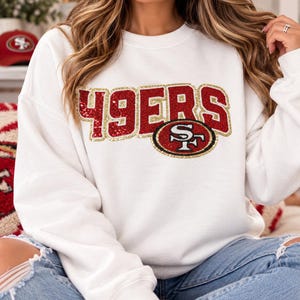 May include: A white long-sleeve crewneck sweatshirt featuring the text 49ERS in red and gold sequins across the chest. The design includes a circular San Francisco 49ers team logo patch also embellished with red, gold, and white sequins.
