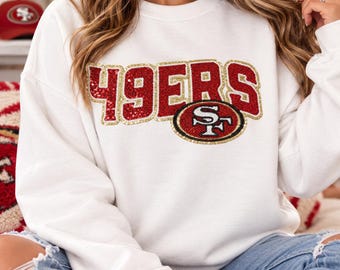 Könnte beinhalten: Ein weißes Langarm-Sweatshirt mit Rundhalsausschnitt und dem Schriftzug 49ERS aus roten und goldenen Pailletten auf der Brust. Das Design enthält einen kreisförmigen Aufnäher mit dem Logo der 49ers, der ebenfalls mit roten, goldenen und weißen Pailletten besetzt ist.