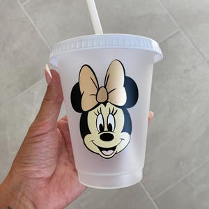 Puede incluir: Un vaso de pl&aacute;stico transparente con tapa y pajita. El vaso presenta un gr&aacute;fico de Minnie Mouse con un lazo dorado y orejas negras. La taza est&aacute; dise&ntilde;ada para bebidas fr&iacute;as.