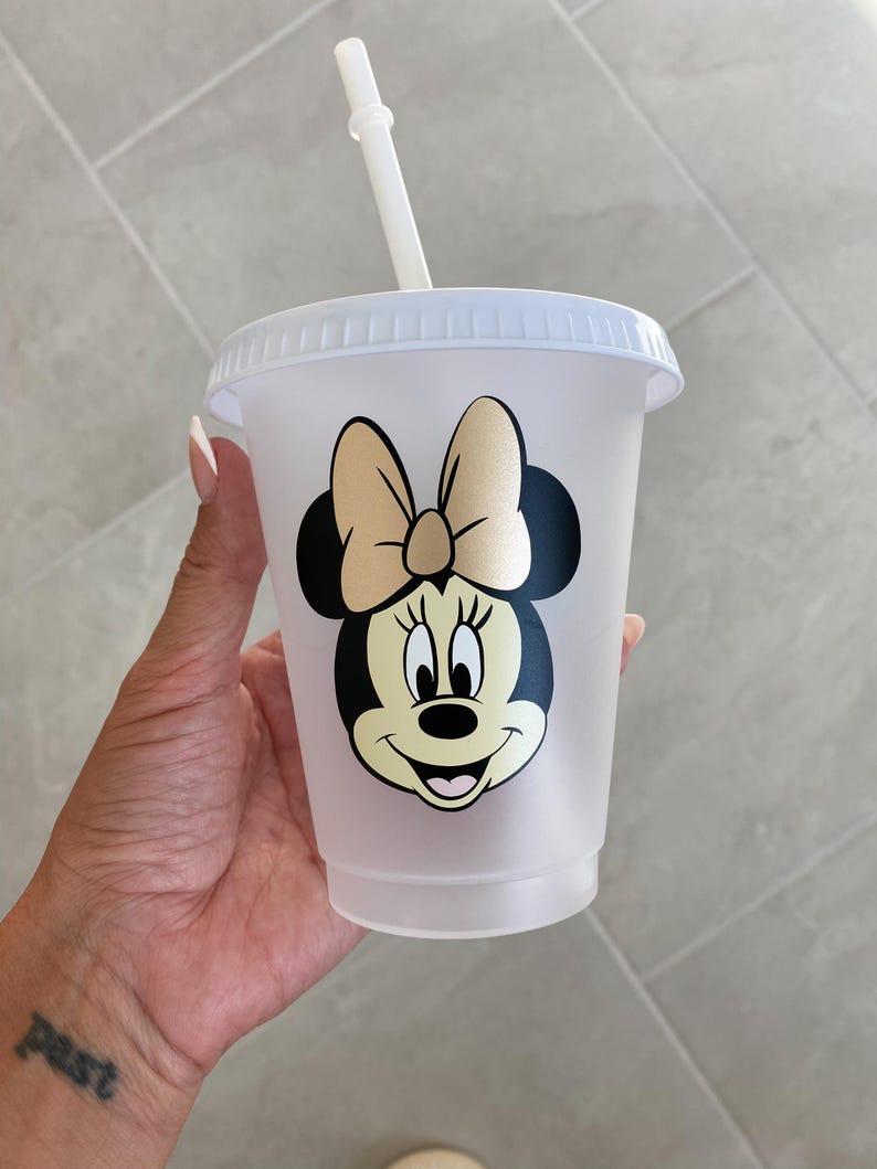 Puede incluir: Un vaso de pl&aacute;stico transparente con tapa y pajita. El vaso presenta un gr&aacute;fico de Minnie Mouse con un lazo dorado y orejas negras. La taza est&aacute; dise&ntilde;ada para bebidas fr&iacute;as.