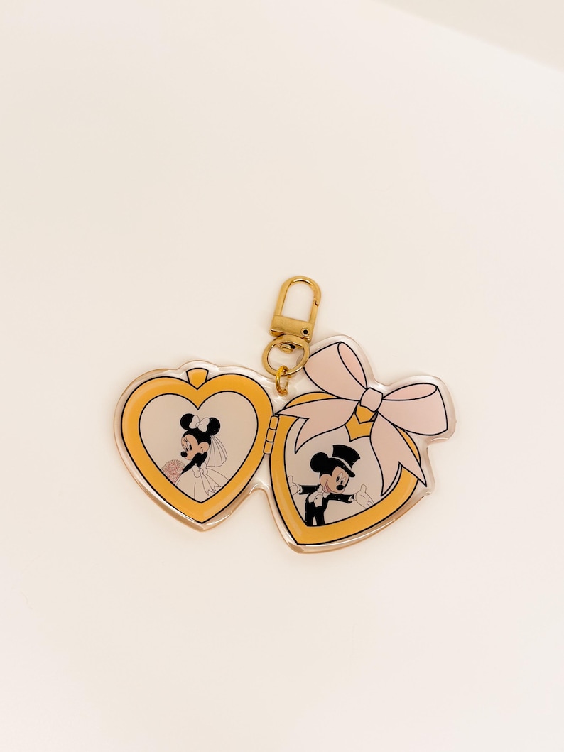 May include: A heart-shaped keychain featuring Mickey and Minnie Mouse in wedding attire. The keychain has a gold-colored clasp and a pink bow. The images of Mickey and Minnie are inside heart-shaped frames.