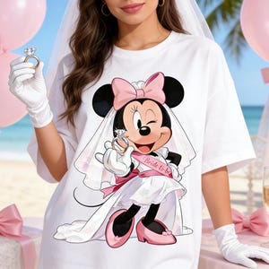 May include: White t-shirt featuring a cartoon Minnie Mouse in a bridal outfit, holding a diamond ring. Minnie wears a veil, pink bow, and a sash that says "Bride to be". Pink balloons and champagne glasses are in the background.
