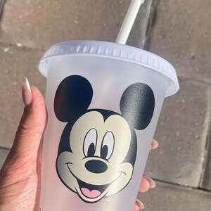 Puede incluir: Un vaso de pl&aacute;stico transparente con tapa y pajita blancas. El vaso presenta un gran gr&aacute;fico de la cara de Mickey Mouse en negro, blanco y rosa. El vaso est&aacute; sostenido por una persona.