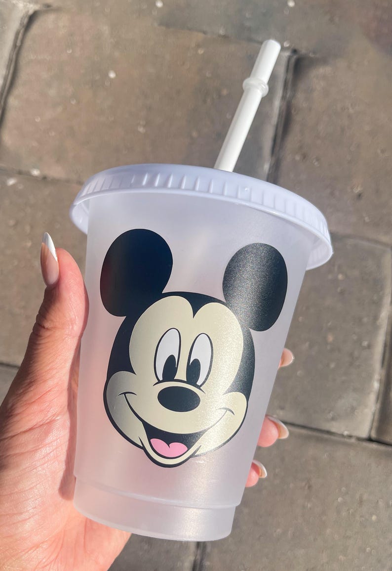 Puede incluir: Un vaso de pl&aacute;stico transparente con tapa y pajita blancas. El vaso presenta un gran gr&aacute;fico de la cara de Mickey Mouse en negro, blanco y rosa. El vaso est&aacute; sostenido por una persona.