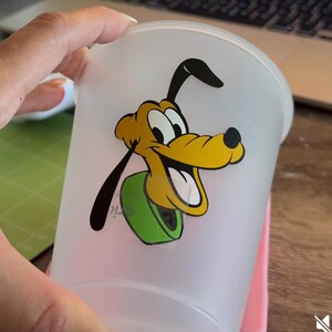 Puede incluir: Un vaso de pl&aacute;stico esmerilado con una imagen de dibujos animados de Pluto, un perro amarillo con orejas negras y un collar verde. El vaso est&aacute; sostenido por una mano sobre un fondo borroso.