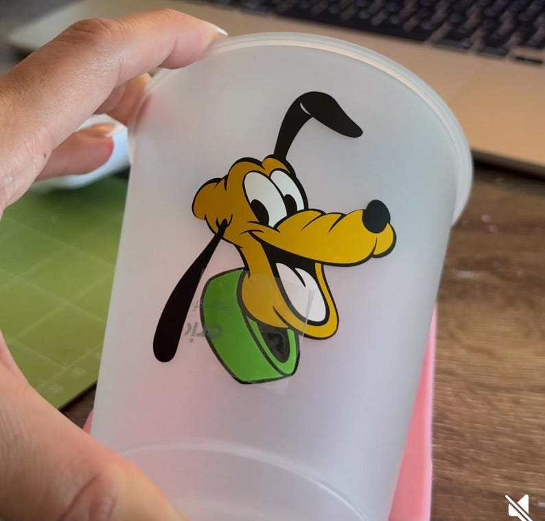Puede incluir: Un vaso de pl&aacute;stico esmerilado con una imagen de dibujos animados de Pluto, un perro amarillo con orejas negras y un collar verde. El vaso est&aacute; sostenido por una mano sobre un fondo borroso.