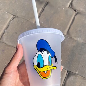 Puede incluir: Un vaso de pl&aacute;stico transparente con tapa y pajita blancas. El vaso presenta un gr&aacute;fico colorido de Pato Donald. El personaje de dibujos animados tiene un sombrero azul, pico amarillo y pajarita negra.