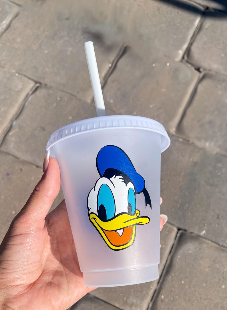Puede incluir: Un vaso de pl&aacute;stico transparente con tapa y pajita blancas. El vaso presenta un gr&aacute;fico colorido de Pato Donald. El personaje de dibujos animados tiene un sombrero azul, pico amarillo y pajarita negra.