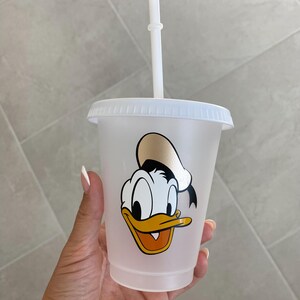 Puede incluir: Un vaso de pl&aacute;stico transparente con tapa y pajita blancas. El vaso presenta una imagen de dibujos animados de Pato Donald. La pajita y la tapa son blancas. La imagen de Pato Donald es en amarillo, naranja, negro y blanco.