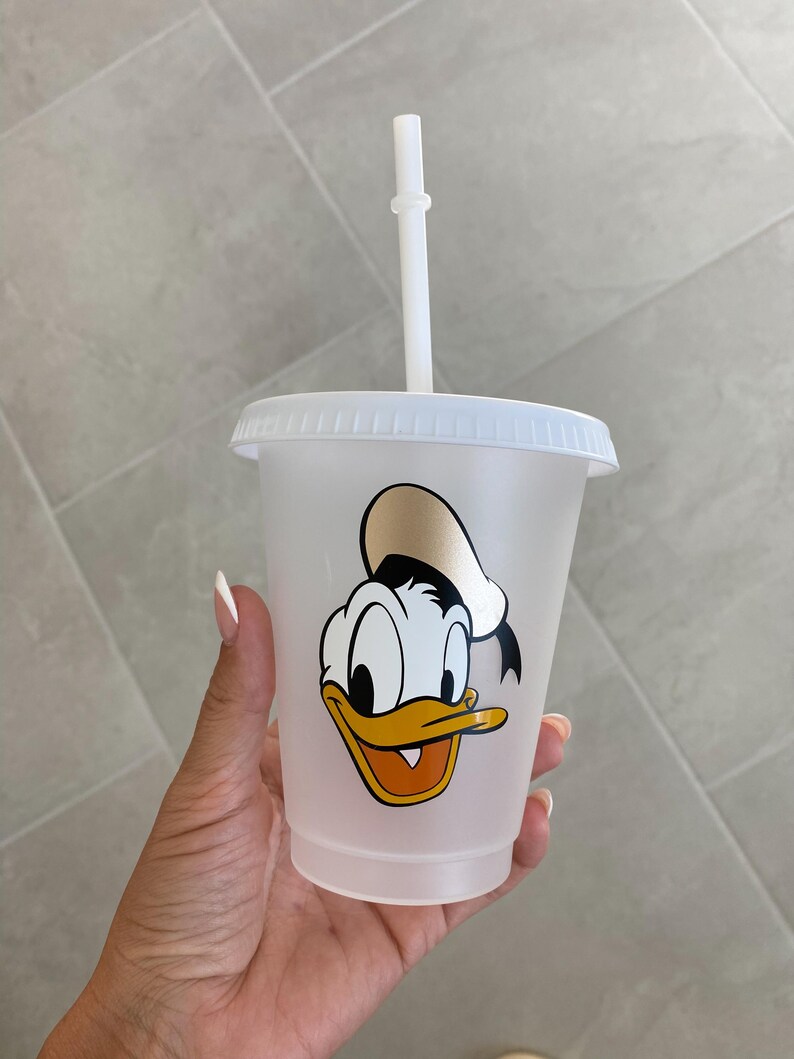 Puede incluir: Un vaso de pl&aacute;stico transparente con tapa y pajita blancas. El vaso presenta una imagen de dibujos animados de Pato Donald. La pajita y la tapa son blancas. La imagen de Pato Donald es en amarillo, naranja, negro y blanco.