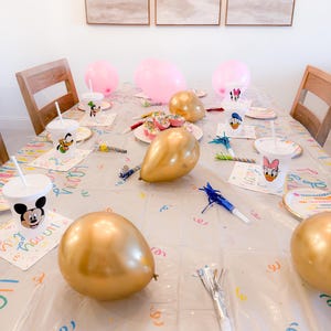 Puede incluir: Una mesa de fiesta decorada con globos rosas y dorados, cupcakes y vasos y platos con personajes. El mantel transparente tiene confeti de colores y servilletas con la inscripci&oacute;n "Happy Birthday". Art&iacute;culos de fiesta y serpentinas.