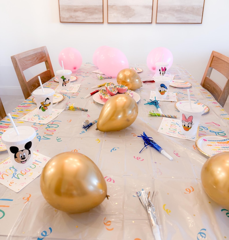 Puede incluir: Una mesa de fiesta decorada con globos rosas y dorados, cupcakes y vasos y platos con personajes. El mantel transparente tiene confeti de colores y servilletas con la inscripci&oacute;n "Happy Birthday". Art&iacute;culos de fiesta y serpentinas.