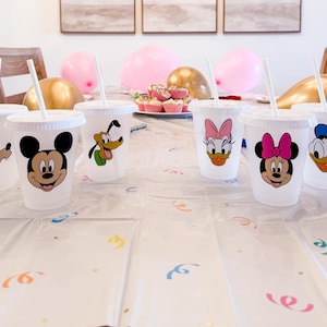 Puede incluir: Una colecci&oacute;n de vasos de pl&aacute;stico transparente con tapas y pajitas blancas, cada uno con un personaje de dibujos animados diferente. Los personajes incluyen Mickey Mouse, Minnie Mouse, Pato Donald, Pata Daisy, Goofy y Pluto. Globos rosas y cupcakes en el fondo.
