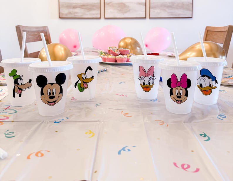 Puede incluir: Una colecci&oacute;n de vasos de pl&aacute;stico transparente con tapas y pajitas blancas, cada uno con un personaje de dibujos animados diferente. Los personajes incluyen Mickey Mouse, Minnie Mouse, Pato Donald, Pata Daisy, Goofy y Pluto. Globos rosas y cupcakes en el fondo.