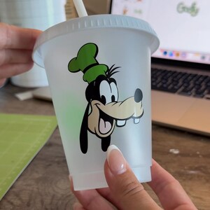 Puede incluir: Un vaso de pl&aacute;stico transparente con tapa y pajita blancas. El vaso presenta una imagen de dibujos animados de Goofy en negro, verde y beige. El dise&ntilde;o est&aacute; centrado en el vaso.
