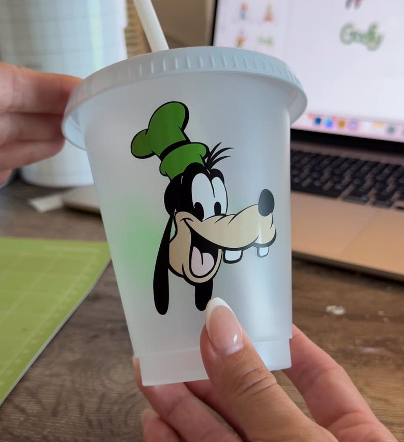 Puede incluir: Un vaso de pl&aacute;stico transparente con tapa y pajita blancas. El vaso presenta una imagen de dibujos animados de Goofy en negro, verde y beige. El dise&ntilde;o est&aacute; centrado en el vaso.