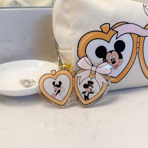 Op de afbeelding: Een hartvormige sleutelhanger met Mickey en Minnie Mouse in trouwkleding. De sleutelhanger is goudkleurig met een transparante acryllaag. Er is een witte strik bevestigd. Een canvas make-uptas met Mickey en Minnie Mouse staat op de achtergrond.