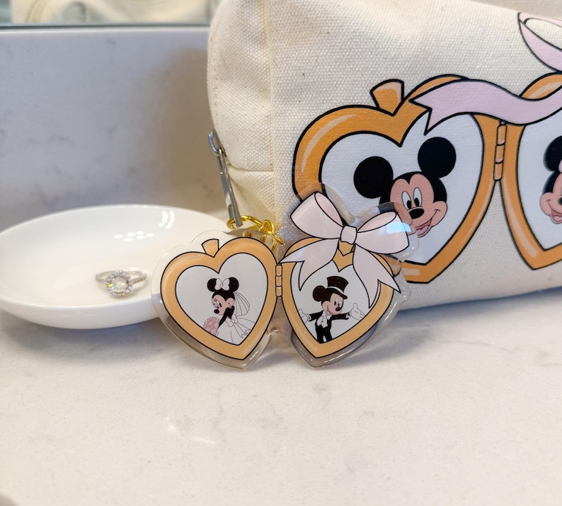 May include: A heart-shaped keychain featuring Mickey and Minnie Mouse in wedding attire. The keychain is gold-toned with a clear acrylic overlay. A white bow is attached. A canvas cosmetic bag with Mickey and Minnie Mouse is in the background.