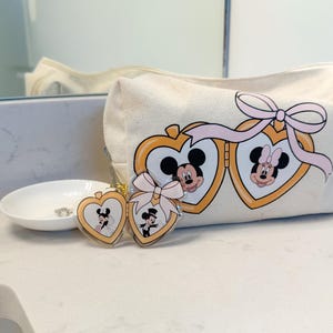 May include: A cream-colored cosmetic bag with a Mickey and Minnie Mouse design. The bag features a pink bow and gold heart-shaped frames. Two matching keychains with Mickey and Minnie in wedding attire are also present.