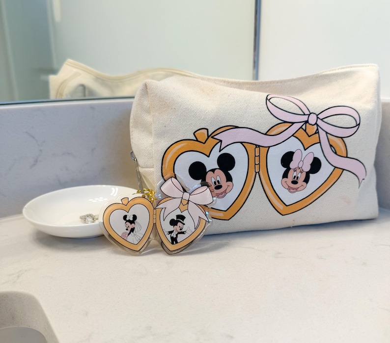 May include: A cream-colored cosmetic bag with a Mickey and Minnie Mouse design. The bag features a pink bow and gold heart-shaped frames. Two matching keychains with Mickey and Minnie in wedding attire are also present.