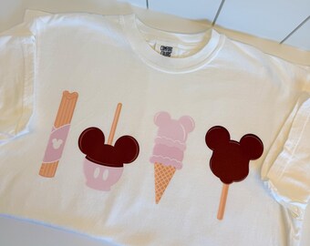 May include: White t-shirt featuring four cartoon food graphics: a churro, a Mickey Mouse-shaped ice cream, a pink ice cream cone, and a Mickey Mouse-shaped popsicle. The shirt has the words "Comfort Colors" printed on the upper back.
