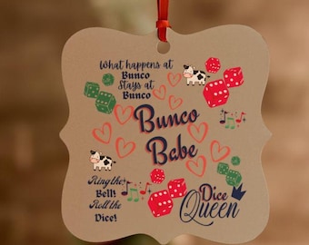 May include: A brown, rectangular, decorative sign with rounded corners and a red ribbon hanging from the top. The sign has a white cow, dice, hearts, and musical notes. The text on the sign reads "What happens at Bunco Stays at Bunco", "Bunco Babe", and "Dice Queen".