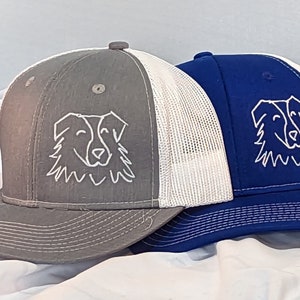Könnte beinhalten: Zwei Trucker-Caps mit einem weißen Hundegesicht, das auf die Vorderseite gestickt ist. Eine Mütze ist grau mit einem weißen Netzrücken, die andere ist blau mit einem weißen Netzrücken.