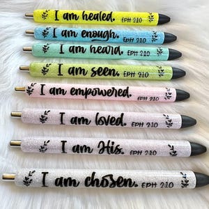 Puede incluir: Una colección de siete bolígrafos con purpurina en varios colores, cada uno con una frase inspiradora en escritura negra. Los bolígrafos incluyen frases como "Soy sanado" y "Soy elegido", con "EPH 2:10" impreso en cada uno.