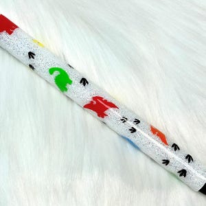 Può includere: Una penna bianca con una finitura scintillante e impronte di dinosauro nere. La penna presenta silhouette di dinosauri colorati, tra cui rosso, verde e giallo.