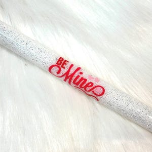 May include: A white glitter pen with the words "BE MINE" in red script. The pen has a Valentine's Day theme with small pink hearts. The pen is on a white, fluffy surface.