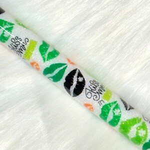 May include: A white glitter tube with green and black lips and the text "Kiss Me More" in black cursive writing.
