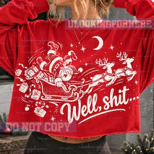 May include: A red long-sleeve cropped sweatshirt featuring a white graphic of Santa Claus falling out of a sleigh pulled by two reindeer. The design includes falling gift boxes, a crescent moon, stars, and the text "Well, shit..." in white script.