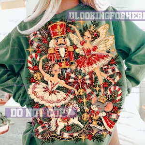 May include: A sage green sweatshirt featuring a festive graphic print of a nutcracker, two ballerinas in red and white tutus, a rocking horse, and a mouse king. The design includes red and black plaid ribbons, candy canes, and holly.