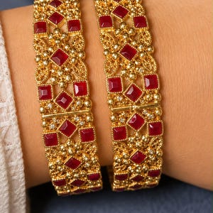 May include: Two gold-toned bangles featuring a repeating pattern of square-cut red stones and intricate metal filigree work. The bangles are constructed with small gold beads and tiny clear accents arranged in a symmetrical, ornate design.
