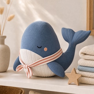 Puede incluir: Un peluche de ballena en color azul oscuro con una barriga de color crema con costuras verticales. La ballena tiene los ojos cerrados, un pequeño detalle de mejilla rosa y lleva una bufanda a rayas crema y rosa atada al cuello.