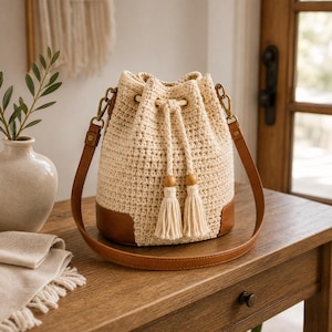 Peut inclure: Un sac seau en crochet de couleur crème avec une base et des garnitures en cuir marron, une fermeture à cordon avec deux pompons assortis et une longue bandoulière en cuir marron fixée par des clips en métal doré sur les côtés.