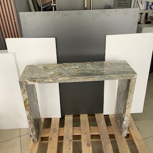 Pu&ograve; includere: Un tavolino da console in marmo verde chiaro con un piano rettangolare e due gambe. Il tavolo &egrave; appoggiato su un pallet di legno.
