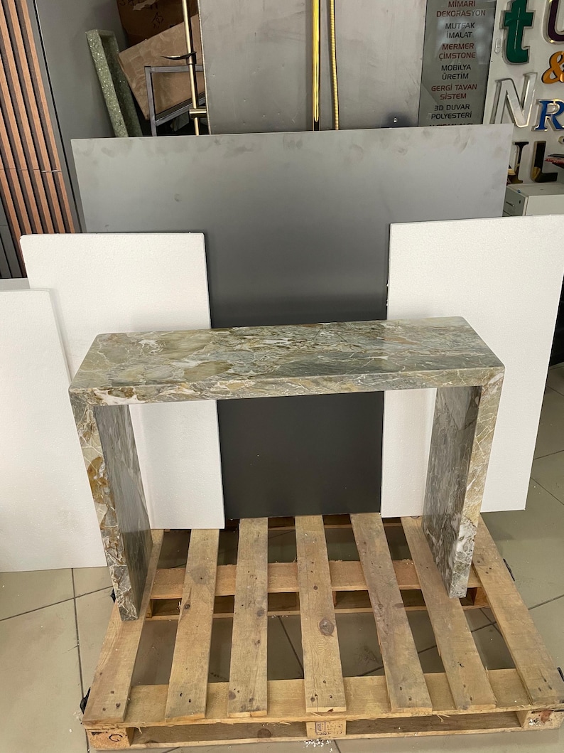 Pu&ograve; includere: Un tavolino da console in marmo verde chiaro con un piano rettangolare e due gambe. Il tavolo &egrave; appoggiato su un pallet di legno.