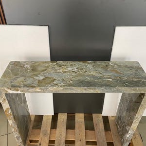 Pu&ograve; includere: Un tavolo consolle in marmo verde e marrone con un piano rettangolare e due gambe. Il tavolo &egrave; appoggiato su un pallet di legno.
