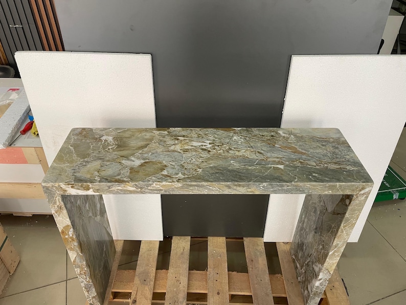 Pu&ograve; includere: Un tavolo consolle in marmo verde e marrone con un piano rettangolare e due gambe. Il tavolo &egrave; appoggiato su un pallet di legno.