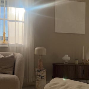 May include: A living room interior with a white couch, a side table with a lamp, and a wooden cabinet. The room is decorated in a minimalist style with neutral colors.