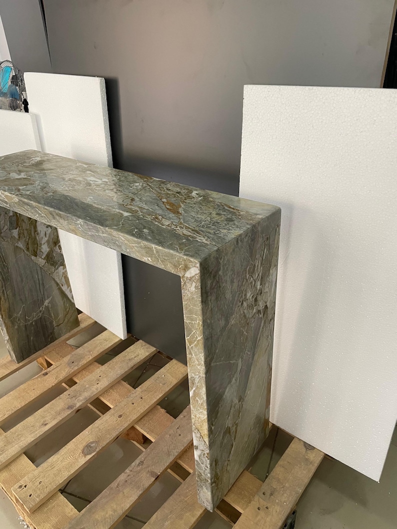 Pu&ograve; includere: Un tavolo consolle in marmo verde con un piano rettangolare e una struttura aperta. Il tavolo &egrave; appoggiato su un pallet di legno.